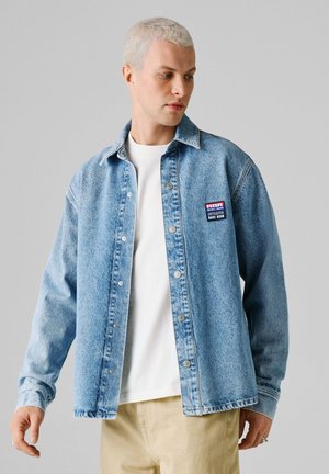 Młody mężczyzna z krótkimi blond włosami, ubrany w jasnoniebieską kurtkę jeansową na białej koszuli i beżowych spodniach, patrzący w lewo.