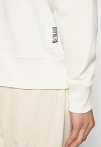 Felpa bianca con polsini e orlo a coste, caratterizzata da un'etichetta verticale nera con logo sul lato sinistro. Tessuto morbido con una texture liscia.