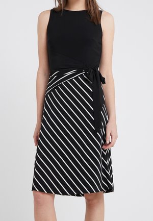 Woman wearing sleeveless black dress with black and white diagonal striped skirt and side tie at waist, standing against plain background.
