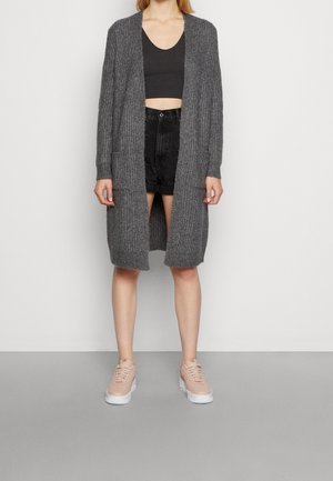 Femme portant un long cardigan gris en maille sur un crop top noir et un short noir, avec des baskets beiges à plateforme, debout sur un sol gris.