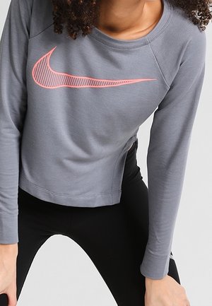 Kobieta ubrana w szarą długą bluzę Nike z różowym logo Swoosh, w połączeniu z czarnymi legginsami, stoi z rękami spoczywającymi na udach.