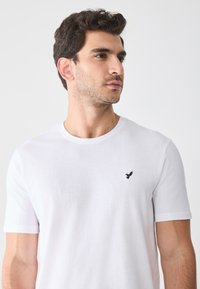 Weißes T-Shirt mit kurzen Ärmeln aus Baumwolle und rundem Halsausschnitt. Verfügt über ein kleines schwarzes Vogel-Logo auf der linken Brust. Glatte Textur.