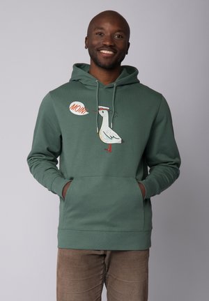 Groene hoodie met een voorzak, met een cartoonzeemeeuw ontwerp en de tekst "MOIN!" in een spraakwolk. Zacht materiaal, relaxte pasvorm.