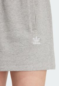 Ljusgrå Adidas-shorts med sidoficka och vit trefoil-logotyp ovanför fållen på bar lår mot en enfärgad bakgrund.