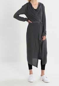 Vestido envolvente gris con escote drapeado, mangas largas y cinturón de lazo. Presenta un detalle de botón en el hombro y una silueta fluida.