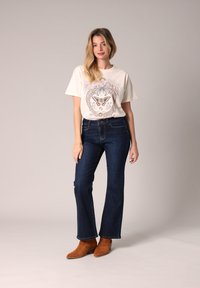 Camiseta gráfica crema con un diseño de mariposa colorido, combinada con jeans acampanados azul oscuro y botines de ante marrón. Fondo sencillo.