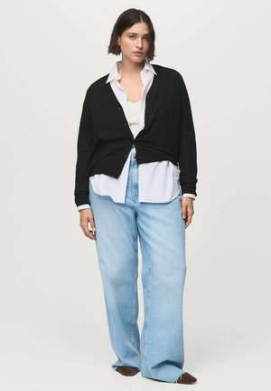 Black cardigan with buttons layered over a white shirt. Light blue wide-leg jeans and brown footwear complete the look. Plain background.