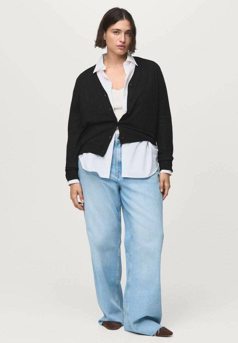 Black cardigan with buttons layered over a white shirt. Light blue wide-leg jeans and brown footwear complete the look. Plain background.