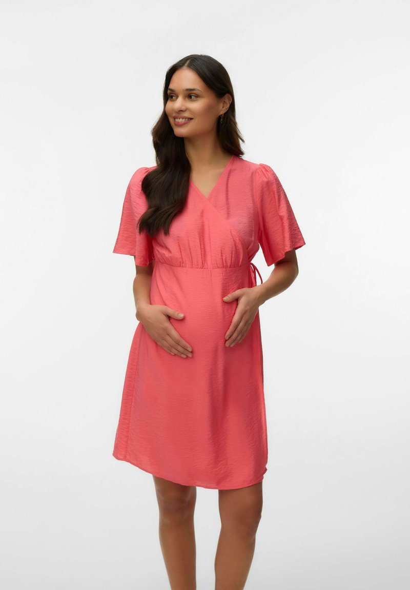 Femme enceinte aux longs cheveux foncés portant une robe corail rose jusqu'aux genoux, tenant doucement son ventre et souriant, sur fond blanc.