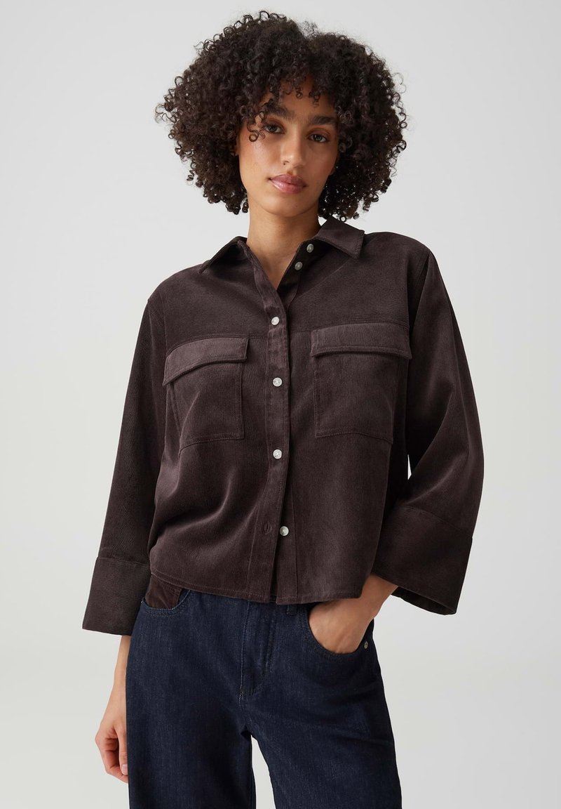 Woman with curly hair wearing a dark brown button-up shirt with two chest pockets and dark blue jeans, posing with one hand in pocket.