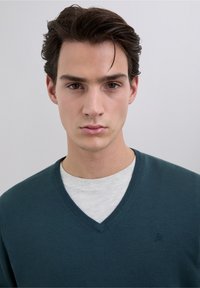 Petrolblauwe v-hals trui gemaakt van zachte stof, gedragen over een lichtgrijs shirt met ronde hals. Heeft een subtiel logo op de linkerborst.