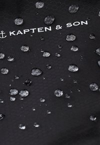 Gouttes d'eau sur un tissu noir à motif hexagonal avec le logo blanc Kapten & Son et un symbole d'ancre.
