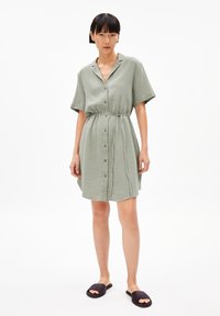 Abito con maniche corte e bottoni di colore verde chiaro, realizzato in tessuto testurizzato con vita fasciante. Abbinato a sandali neri a ciabatta.