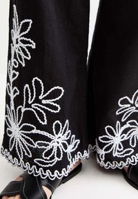Svarta vida byxor med vit blomsterbroderi och vågkant. Bärs med svarta sandaler med ett korsat remdesign.