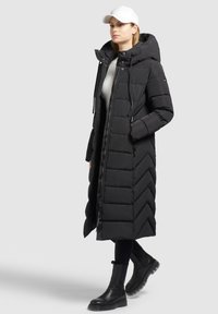 Sort lang pufferjakke med chevronmønster, lynlås foran og hætte. Båret over en lys sweater og parret med sorte knæhøje støvler. Hvid kasket.