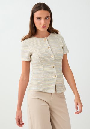 Jeune femme aux longs cheveux bruns portant un haut texturé à manches courtes boutonné et un pantalon beige sur un fond blanc uni.