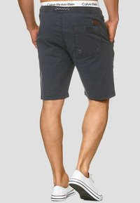 Shorts gris foncé avec une texture en denim, comprenant une poche arrière avec fermeture par bouton, une ceinture élastique et un détail de marque subtil sur la ceinture.