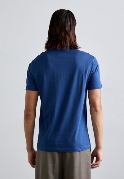Temno modra bombažna majica s krojem za ovratnik in kratkimi rokavi, gladka tekstura, vidni šivi, nosi se z svetlobrunnimi hlačami.