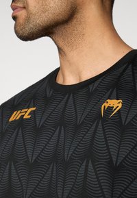 Μαύρο αθλητικό μπλουζάκι με υφή κυματιστού μοτίβου, πορτοκαλί λογότυπο UFC και αντίθετη πορτοκαλί λεπτομέρε ια κοντά στον λαιμό.