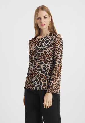 Kobieta o prostych blond włosach, ubrana w długi rękaw w panterkę i czarne spodnie, stojąca na tle jednolitego białego tła.