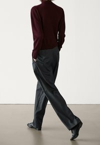 Person, die mit einem weinroten Pullover, einer dunkel gestreiften, weiten Hose und schwarzen spitzen Schuhen davonläuft, mit den Händen in den Taschen.