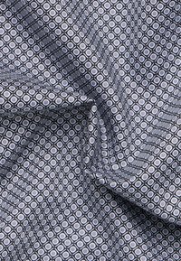 Tessuto fantasia con un design geometrico di piccoli quadrati in blu navy e grigio chiaro, con una superficie liscia e una leggera lucentezza.