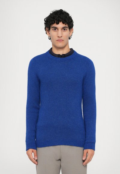 Pull bleu en tissu doux et texturé. Il a un col rond et des manches longues, cintrées aux poignets. Porté sur une chemise à col.