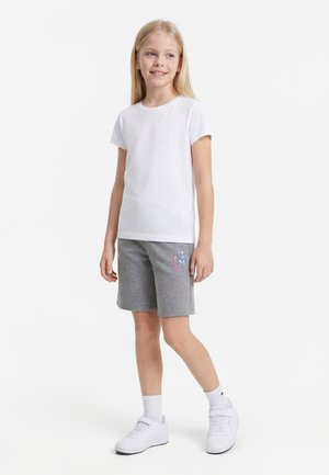 Fille souriante debout, portant un T-shirt blanc à manches courtes, un short gris avec un logo coloré, des chaussettes blanches et des baskets blanches sur un fond uni.