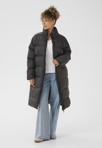 Cappotto imbottito nero lungo con colletto alto, dotato di chiusura a zip e maniche ampie. Indossato sopra una maglietta bianca e jeans azzurri chiari.