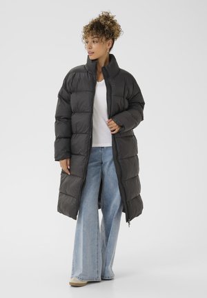 Ilgas, paminkštintas juodas pūkinis paltas su aukštu apykakle, su užtrauktuku ir plačiomis rankovėmis. Dėvimas virš balto marškinėlio ir šviesiai mėlynų džinsų.