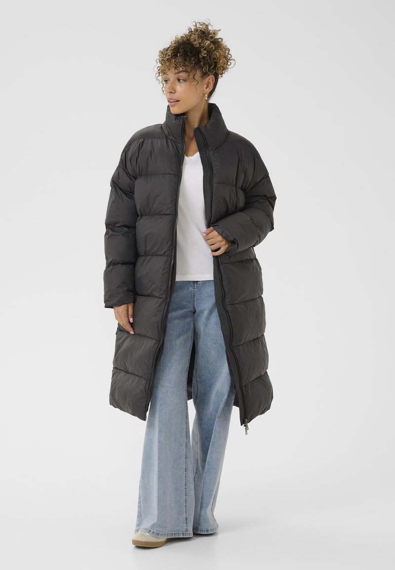 Cappotto imbottito nero lungo con colletto alto, dotato di chiusura a zip e maniche ampie. Indossato sopra una maglietta bianca e jeans azzurri chiari.