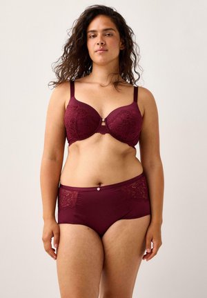 Kvinde med krøllet hår iført matchende burgundy blondebh og højtaljede trusser med blondedetaljer, der står imod en ensfarvet baggrund.