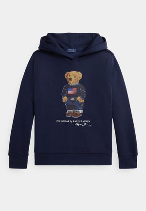 Marineblauwe hoodie met een afbeelding van een teddybeer die een trui met Amerikaanse vlag draagt, een spijkerbroek en schoenen, met daaronder de tekst "Polo Bear by Ralph Lauren".