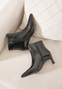 Bottes de cheville en cuir verni noir avec un bout pointu et un petit talon carré. Équipées d'une fermeture éclair sur le côté pour un enfilage facile. Fini glossy texturé.