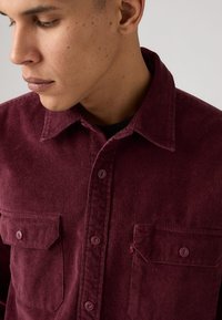 Bourgogne kordflortskjorta med klassisk krage, två bröstfickor och knäppningar; har en texturerad yta och stickade detaljer.