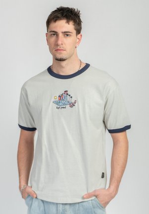 Młody mężczyzna o krótkich ciemnych włosach, ubrany w jasnoszarą koszulkę z granatowym obszyciem i kolorowym nadrukiem przy basenie, stojący z rękami w kieszeniach.