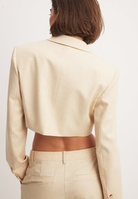 Beige kort blazermed långa ärmar, notchkrage och slät tygkänsla. Har knappdetaljer på ärmarna och rena linjer.