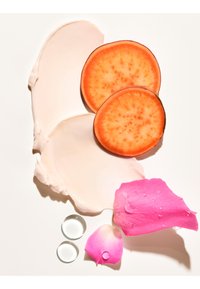 Zwei orangefarbene Süßkartoffelscheiben, cremige weiße Lotion, ein rosafarbenes Rosenblatt mit Tropfen und zwei durchsichtige Kugeln auf weißem Hintergrund.