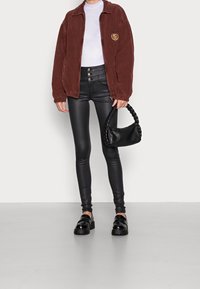 Veste en velours côtelé marron avec un logo, chemise blanche, leggings en faux cuir noir, sac à main noir tressé, et mocassins noirs épais.