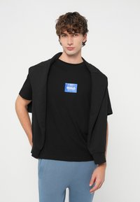 Juoda marškinėliai su mėlynu kvadratu ir užrašu „HUGO“, dėvimi su juodu megztiniu, užmestu per pečius, derinami su šviesiai mėlynomis kelnėmis.