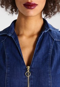 Denimklänning med dragkedja fram och krage, i en djupblå färg och mjuk textur. Guld detaljer framhäver dragkedjeelementet.