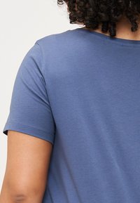 Una vista en primer plano de la espalda de una persona, con las mangas cortas de una camiseta azul, y con el cabello rizado visible por encima del cuello.