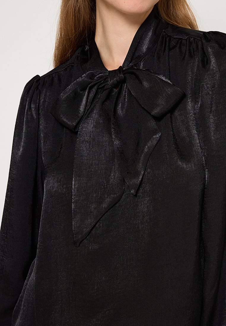 Blusa negra hecha de una tela suave y brillante. Presenta un cuello alto con un gran lazo y hombros fruncidos. Mangas largas con textura suave.