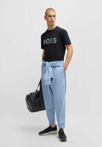 T-shirt en coton noir avec logo "BOSS", associé à un pantalon de survêtement bleu clair. Le modèle tient un sac de sport noir et porte des baskets navy.
