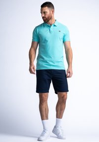 Tyrkysové polo tričko s límečkem, námořnické šortky a bílé tenisky. Má krátké rukávy a malé logo na hrudi.