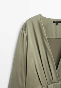Blouse vert olive clair à manches longues avec un col en V profond et des plis de tissu lâches à la taille, vue depuis le côté supérieur gauche.
