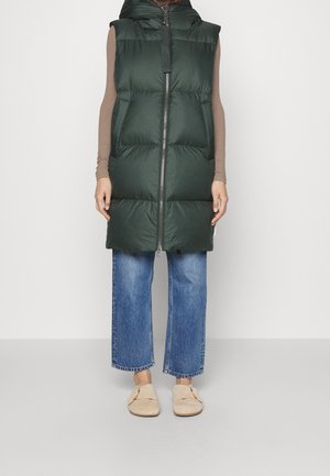Personne portant un gilet sans manches matelassé vert foncé par-dessus un haut beige à manches longues, un jean bleu raccourci et des chaussures beiges à enfiler, debout contre un mur blanc.