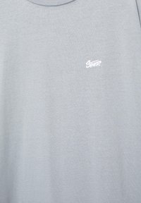 Jasnoszara koszulka z okrągłym dekoltem, z małym białym stylizowanym logo "Swtp." na lewej piersi.