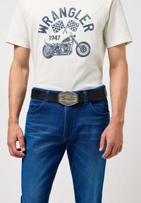 Zwarte leren riem met een zilverkleurige metalen Wrangler-buckle, gedragen over blauwe denim jeans en een crème T-shirt met een motorfietsafbeelding.