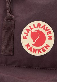 Bordowy plecak z tkaniny z okrągłą kremową naszywką z czerwonym logo lisa i napisem "FJÄLLRÄVEN KÅNKEN" wytłuszczonym czerwonym drukiem.